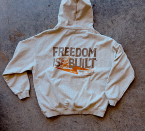 Beige hoodie with 'Freedom is Built' text on a concrete surface