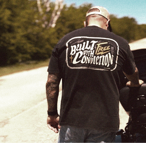 Man wearing a black t-shirt with 'Built with Conviction' and the Free logo walking on a road. Brand: Free is Life