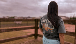 Person wearing a shirt with text standing in front of a wooden fence and open field.