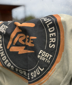 Tan shirt with closeup of graphic on back hanging on wooden fence.