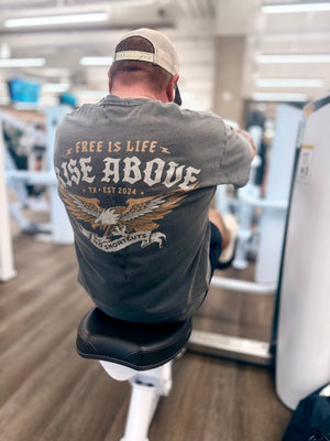 Man wearing a gray tee with 'Free is Life, Rise Above' text in a gym setting.
